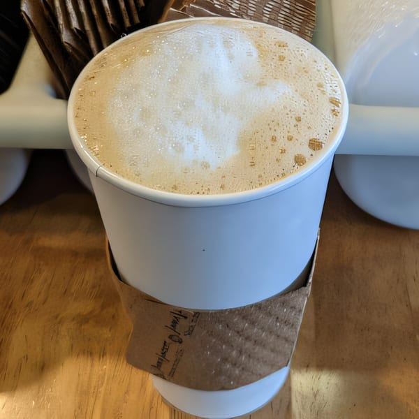 A white paper cup of frothy latte with a brown sleeve sits on a wooden surface.