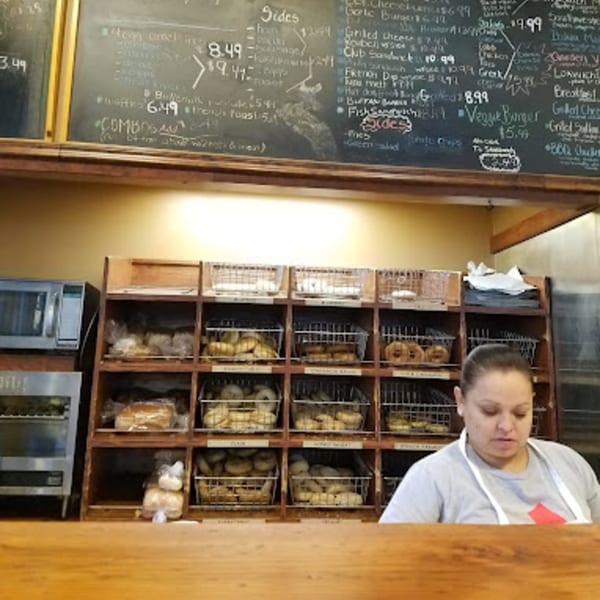 A Wasatch Bagel and Grill employee stands behind a counter with shelves of bagels and a menu board.