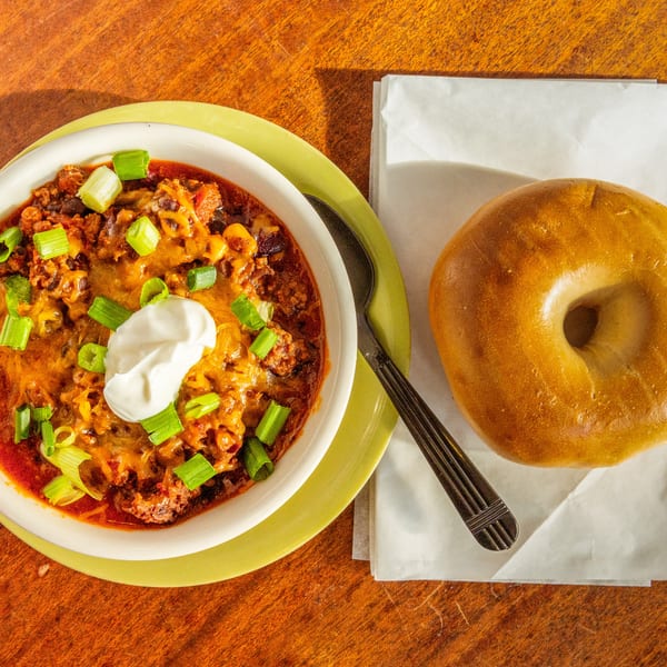 A bowl of chili with cheese, sour cream, and green onions, next to a plain bagel.