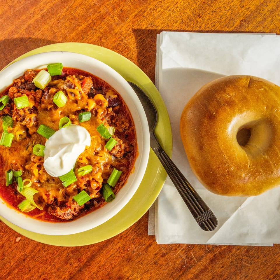 A bowl of chili with cheese, sour cream, and green onions, next to a plain bagel.