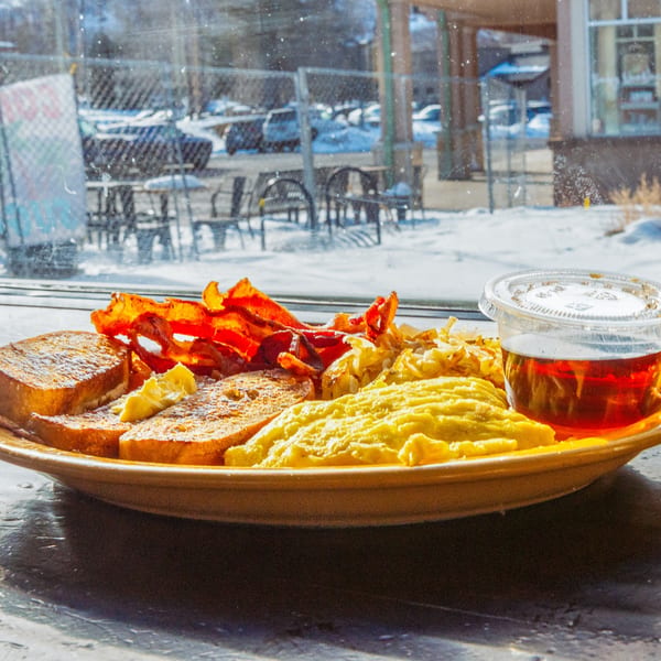 Breakfast plate with French toast, bacon, scrambled eggs, hash browns, and syrup, by a snowy window.