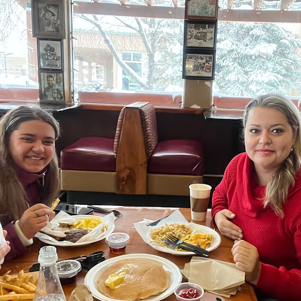Two women smiling at a table with pancakes, fries, and other food at Wasatch Bagel and Grill.