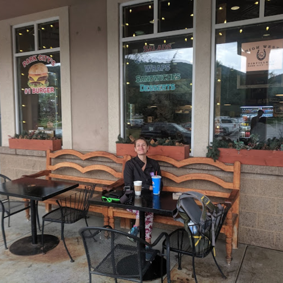 A woman sits at an outdoor table with drinks in front of Wasatch Bagel and Grill.